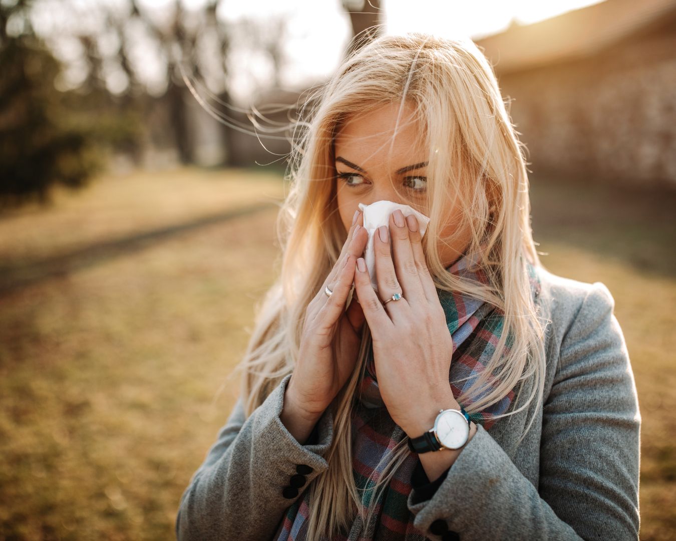 Young blonde woman outside during the fall blowing her nose with a tissue.