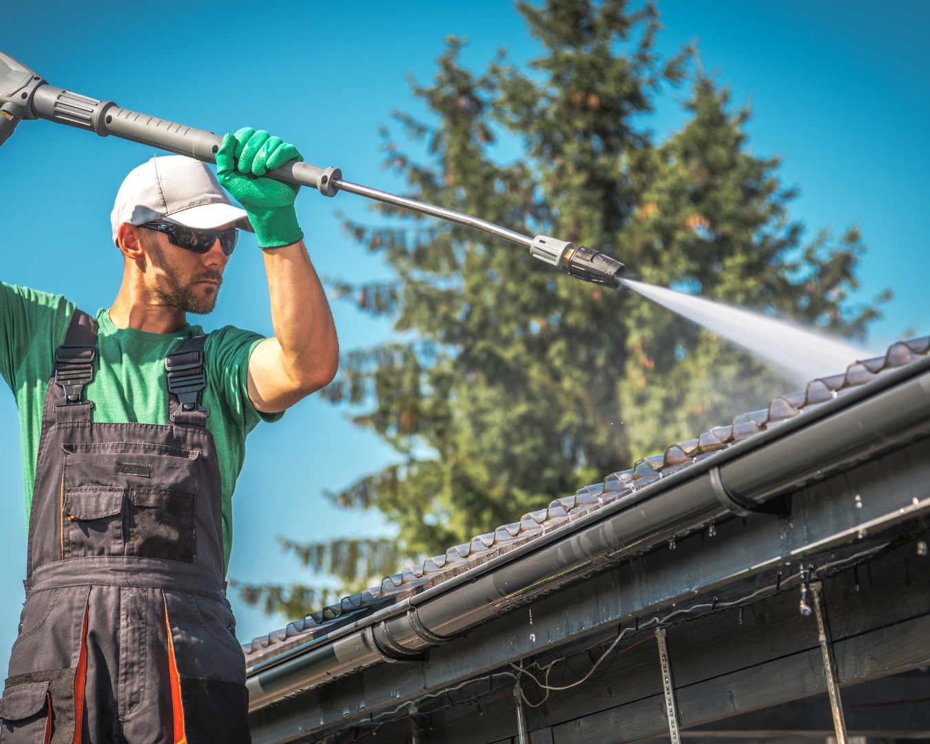 Man in black overalls and a green t-shirt pressure washing a roof, holding the pressure washer overhead.