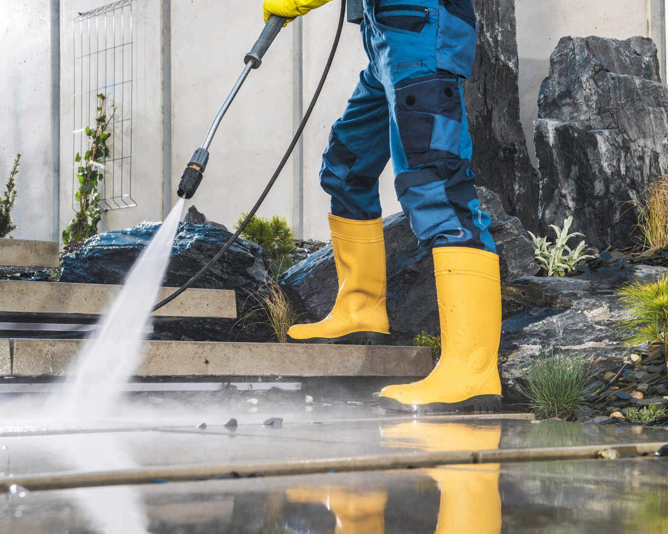 Person pressure washing outdoor concrete steps wearing blue overalls and yellow boots.