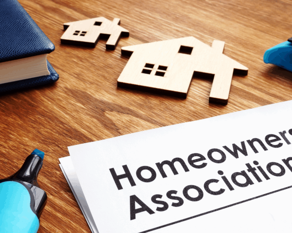 Documents on desk labeled "Homeowners Association" surrounded by pens, books, and wooden figurines of houses.