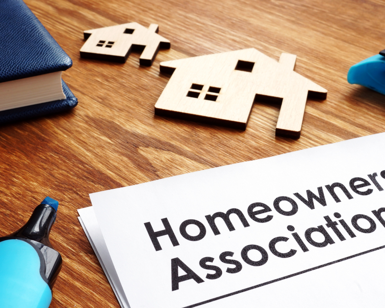 Documents on desk labeled "Homeowners Association" surrounded by pens, books, and wooden figurines of houses.