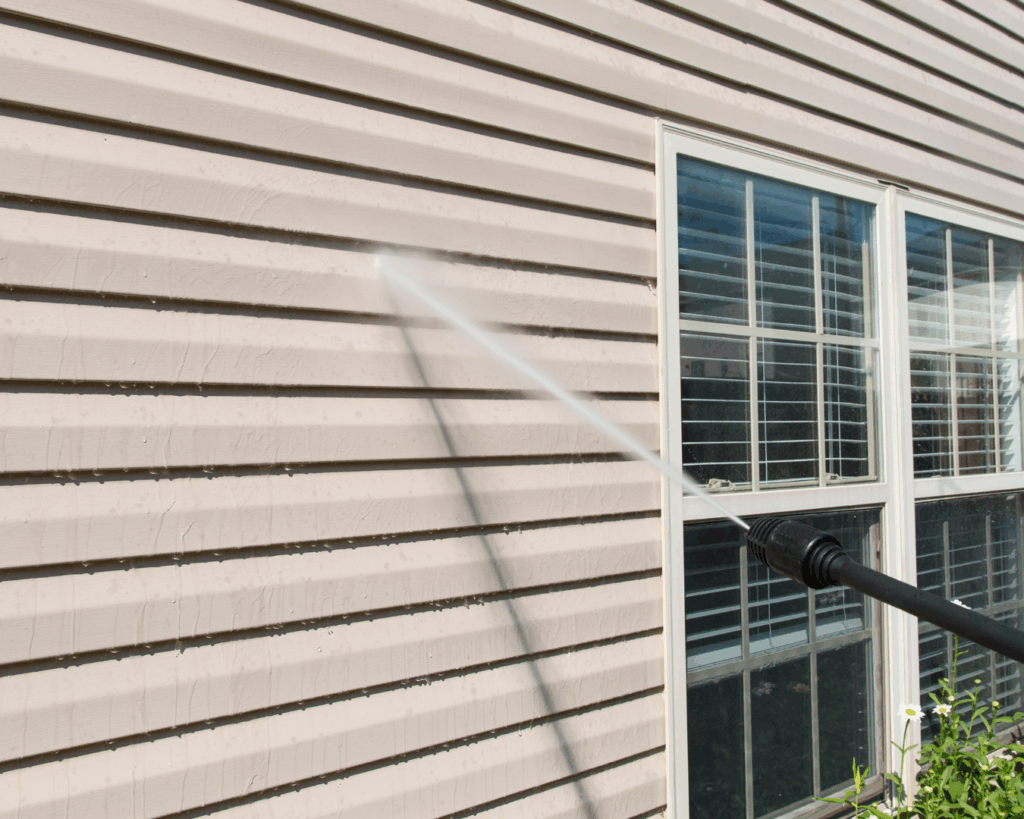 A power washer sprays water onto the beige vinyl siding of a house near a window, removing dirt and grime to restore a clean surface.