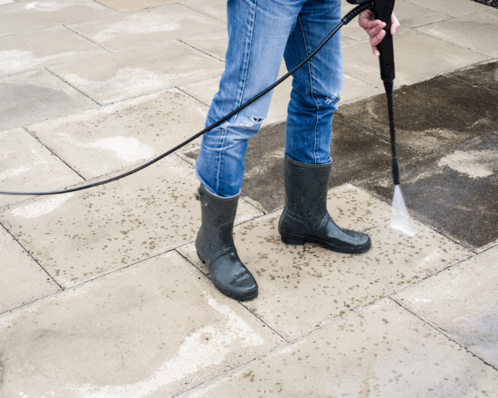 Person wearing jeans and rubber boots using a power washer to clean dirt and grime from outdoor patio pavers.