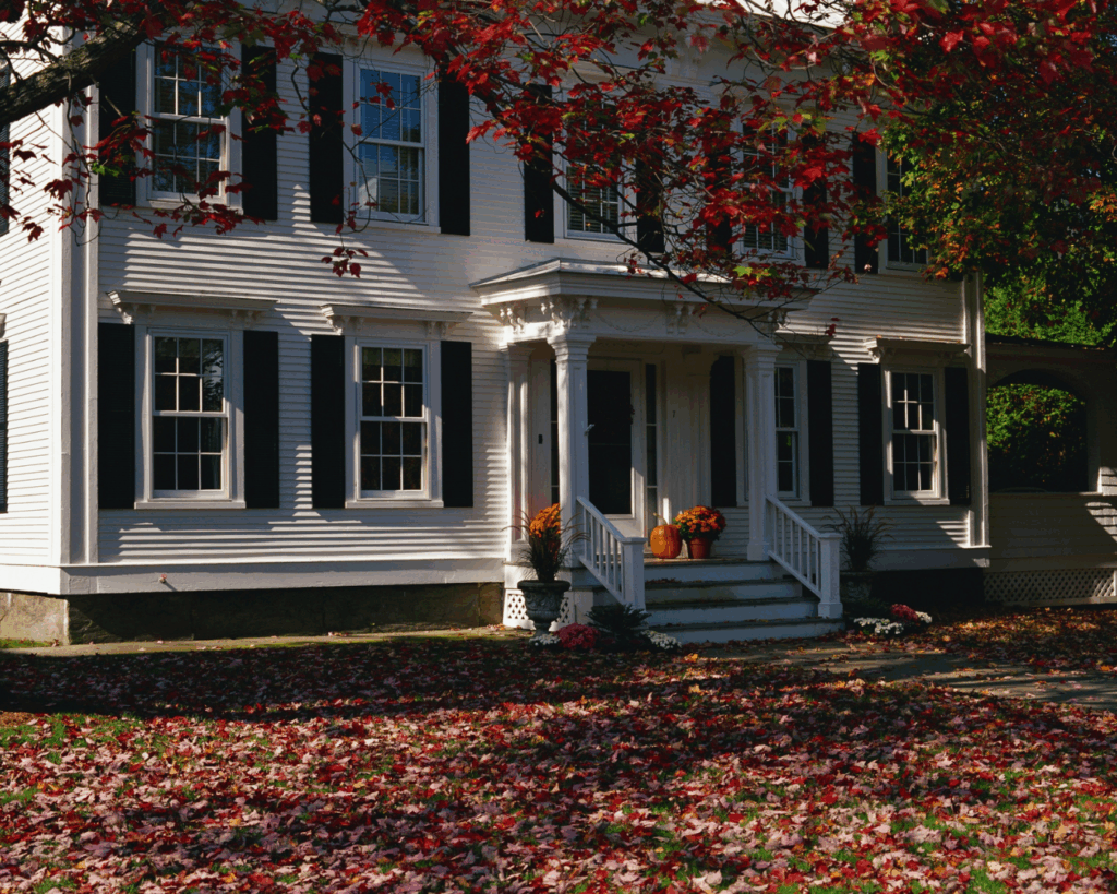 The image shows a charming home surrounded by colorful autumn leaves, with the vibrant reds of the tree branches in the foreground. The front porch of the house features seasonal decorations, including potted plants and pumpkins. The exterior of the house is a classic white with dark shutters, giving it a crisp and timeless appearance. The leaves scattered around the yard create a cozy, fall atmosphere.
