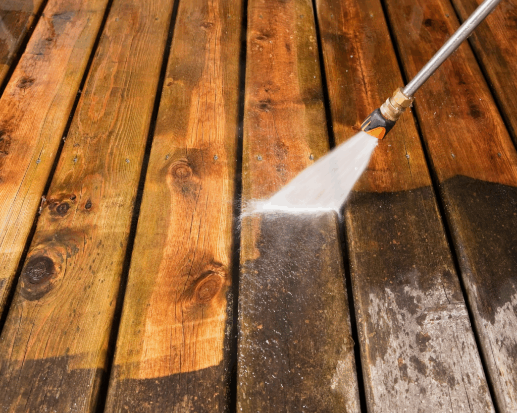 Pressure washer cleaning wooden deck boards. The water stream is visibly cleaning the wood, revealing a brighter and cleaner section on the right, while the left part still shows dirt and discoloration. The contrast between the cleaned and dirty wood highlights the effectiveness of pressure washing for restoring the appearance of outdoor surfaces.