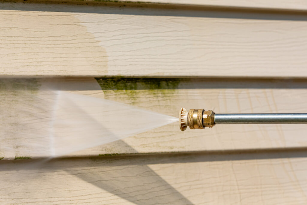 Close-up of a pressure washer cleaning green algae from beige house siding, showing a clear contrast between the cleaned and dirty areas.