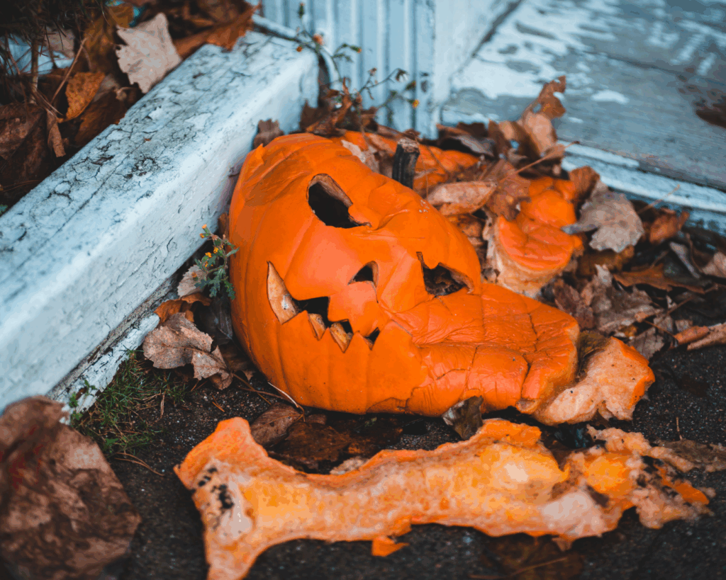 A rotting carved pumpkin collapsing on a wooden porch surrounded by fallen leaves, showing signs of decay and moisture damage.