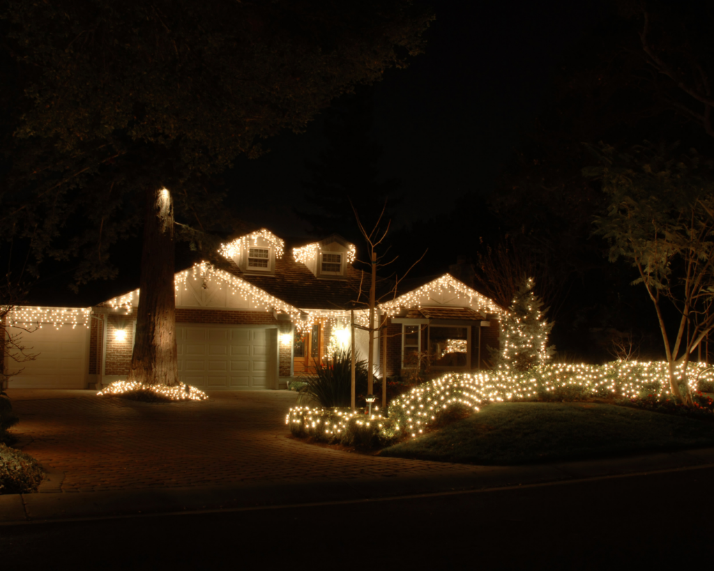 Home decorated with elegant white Christmas lights along the roofline, trees, and landscaping, glowing warmly at night.